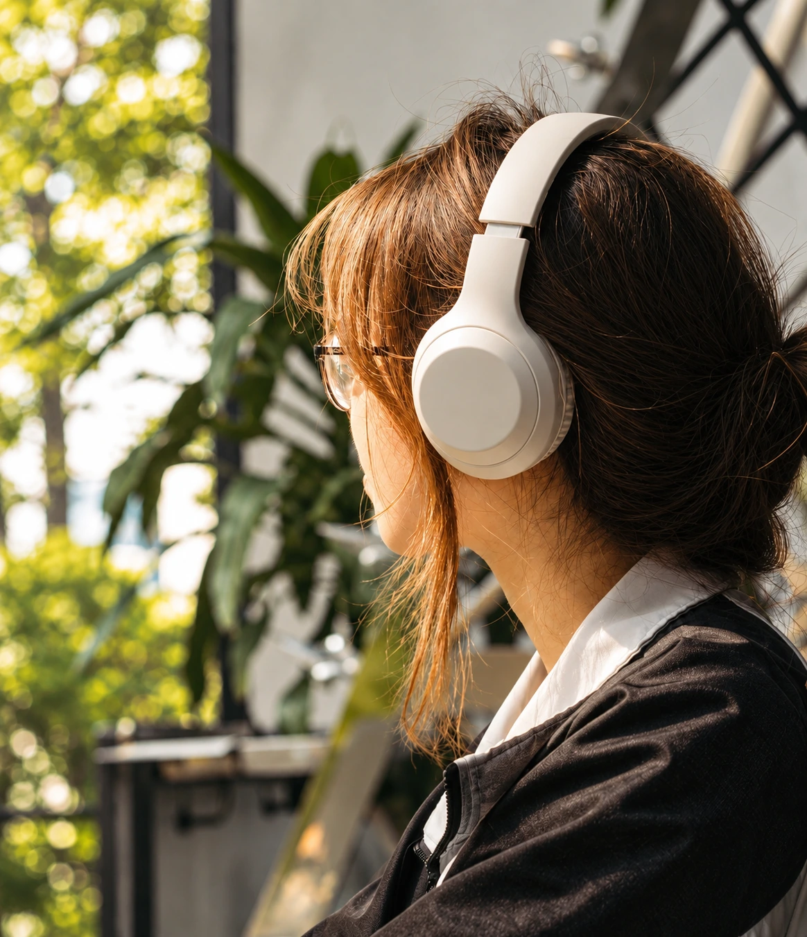 Woman with glasses wearing cream headphones, seen from behind, relaxing near green plants, representing neuro-acoustic biological support for workforce resilience and burnout prevention